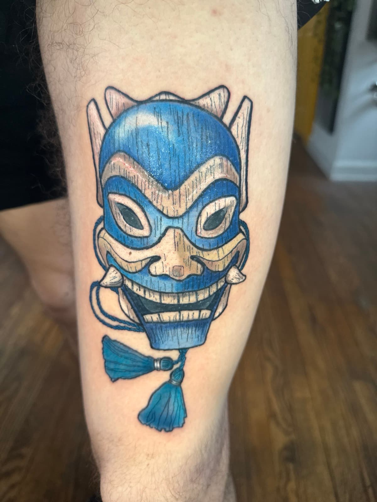 Blue Spirit mask tattoo from Avatar The Last Airbender on calf — wooden mask with blue paint, fangs, and a hanging tassel.