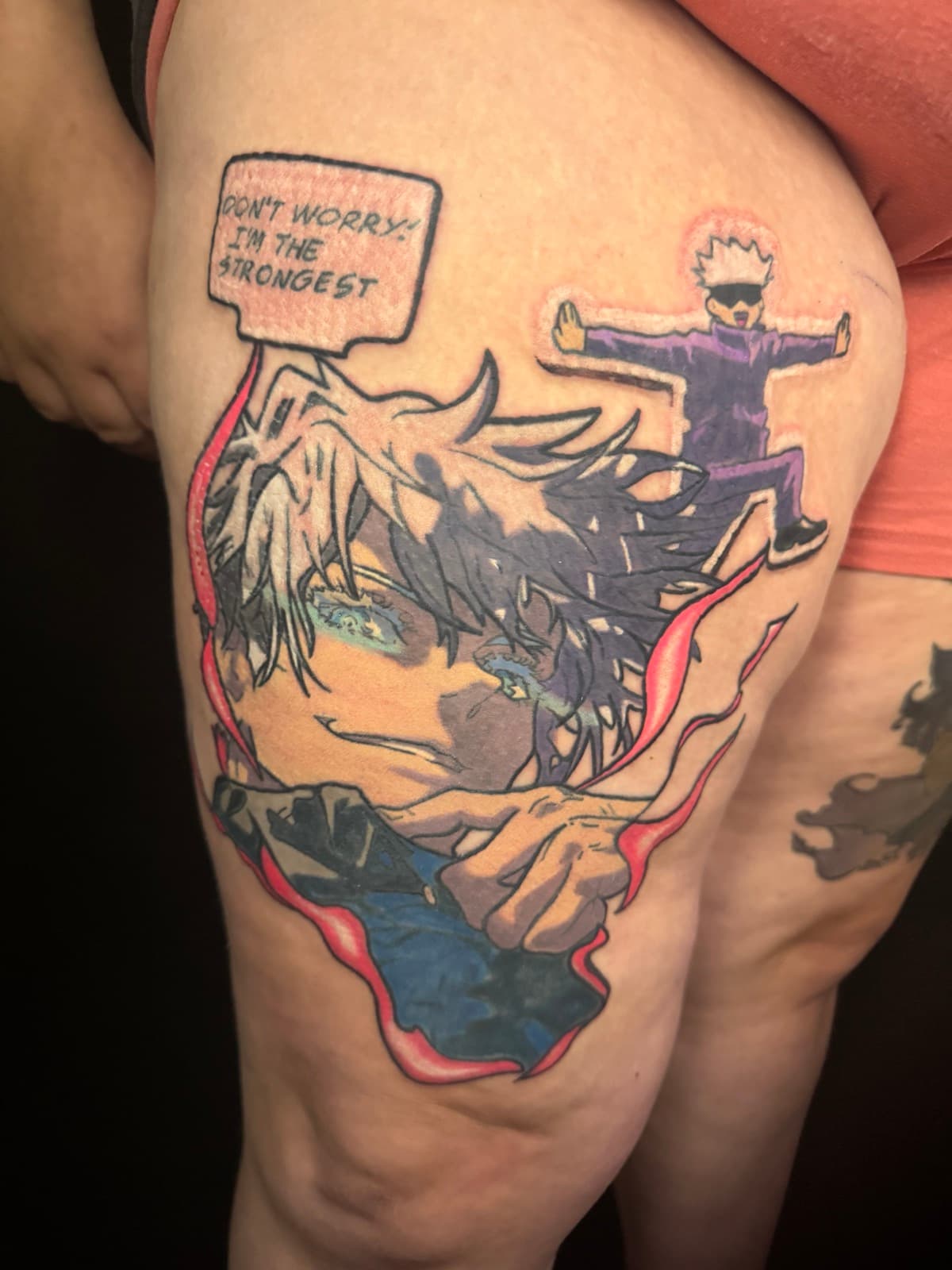Close-up of Jujutsu Kaisen Gojo Satoru tattoo on thigh, fresh ink, with chibi Gojo and "Don't Worry I'm The Strongest" speech bubble.