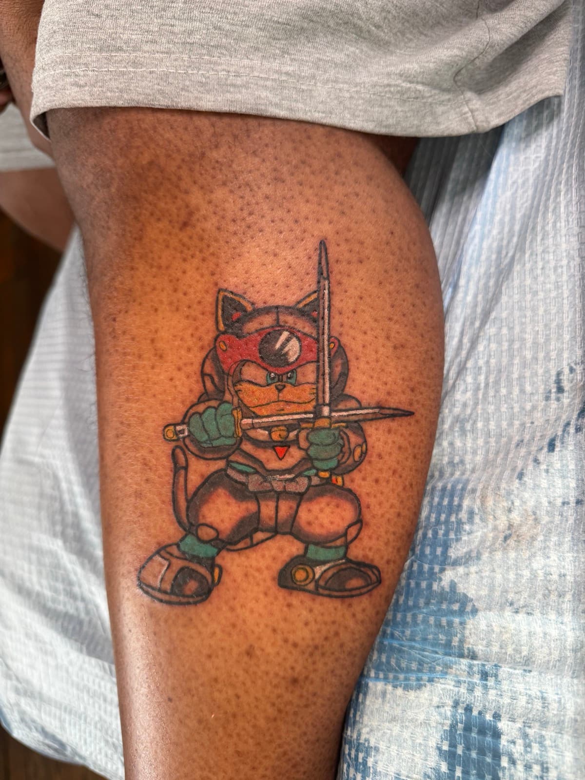 Samurai Pizza Cats tattoo on the leg — orange and brown cat in armor with red eye-mask holding two katana swords.