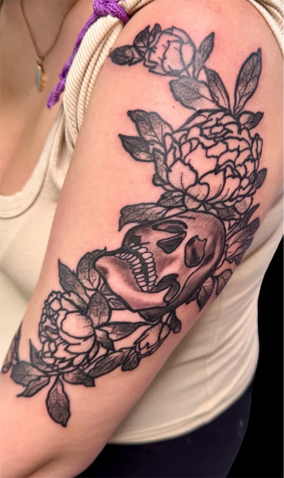 Black and grey upper-arm tattoo of a screaming skull surrounded by detailed peony flowers and leaves.