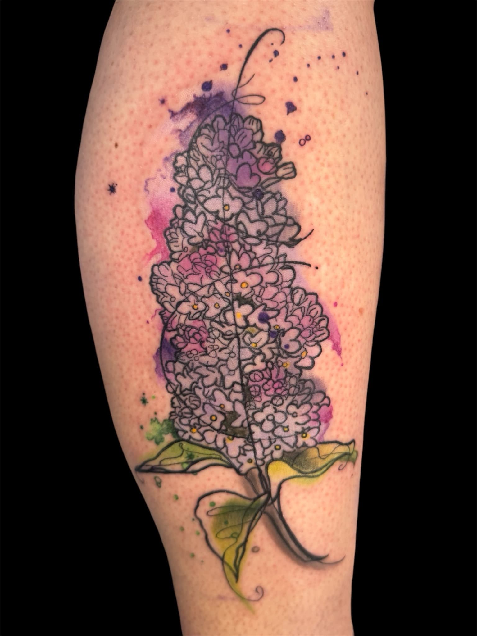 Watercolor lilac tattoo on calf — line-drawn lilac flower cluster with purple and pink watercolor splash and green leaves.
