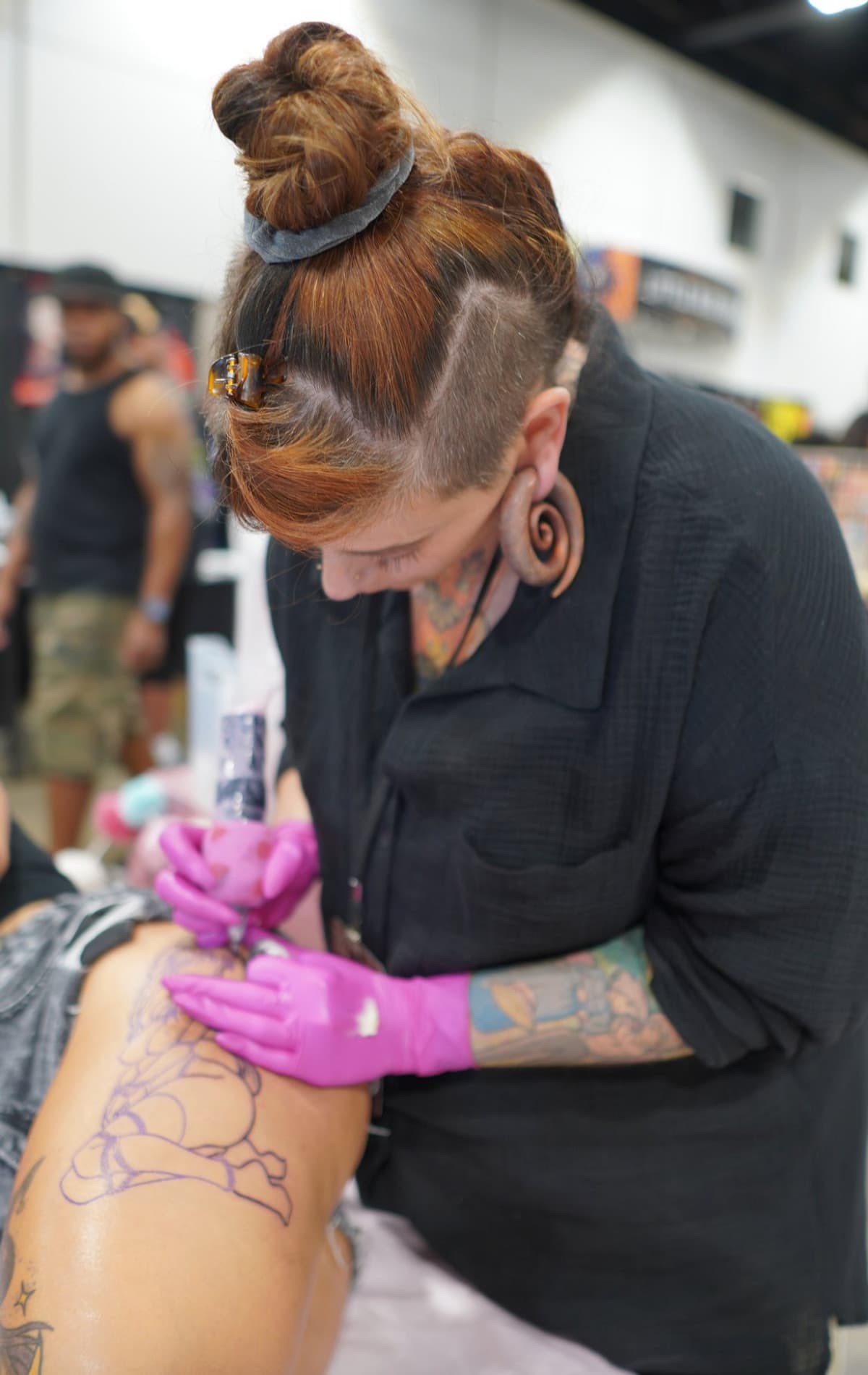 Paula in profile working on a floral tattoo at a convention — concentrated expression, pink gloves, purple stencil lines visible on client's thigh.