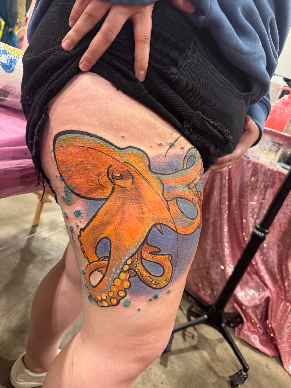 Vibrant orange and teal watercolor octopus tattoo on thigh, photographed at a tattoo convention with pink and blue booth backdrop.