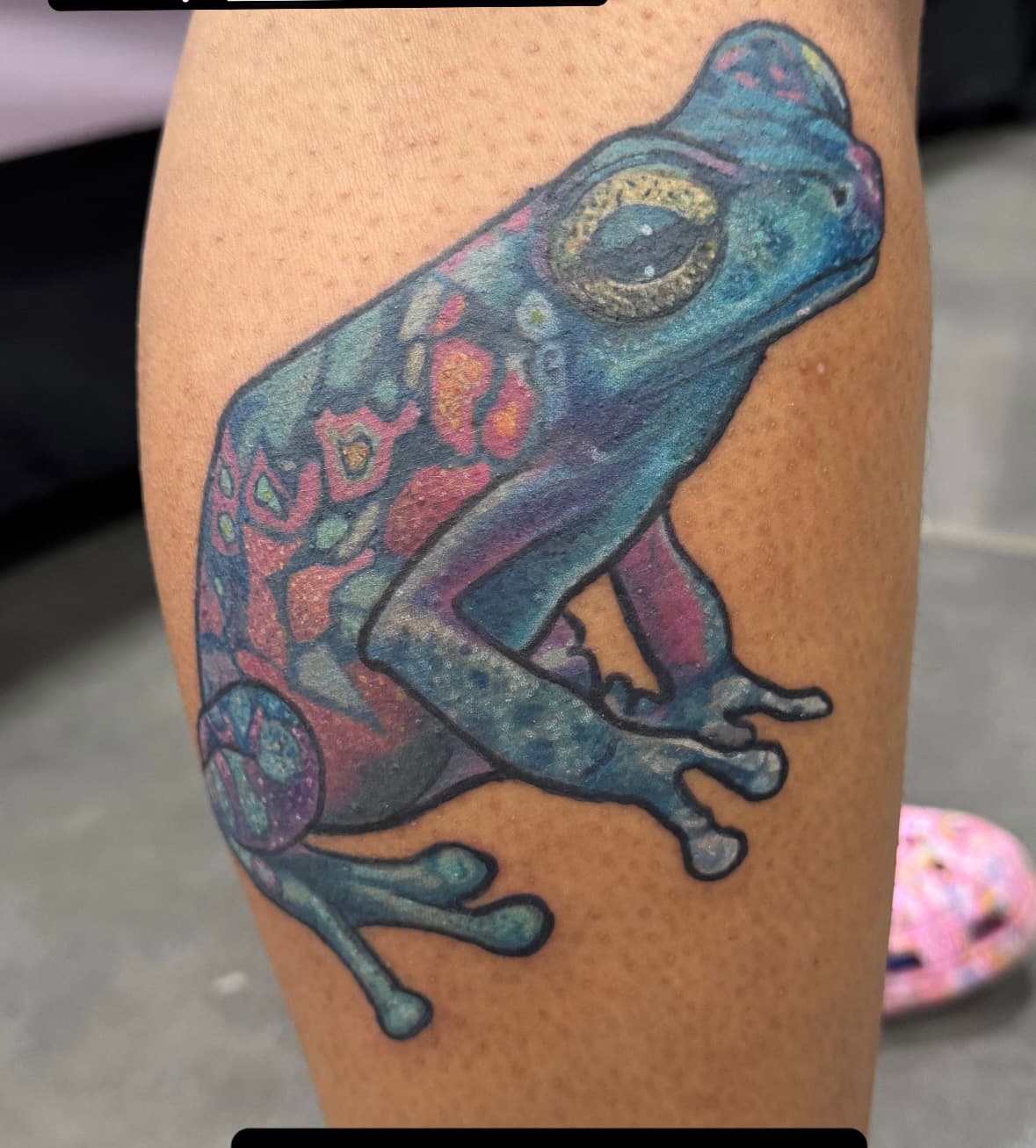 Colorful frog tattoo on calf — blue body with pink, red, and orange patterned back markings and large gold eyes.