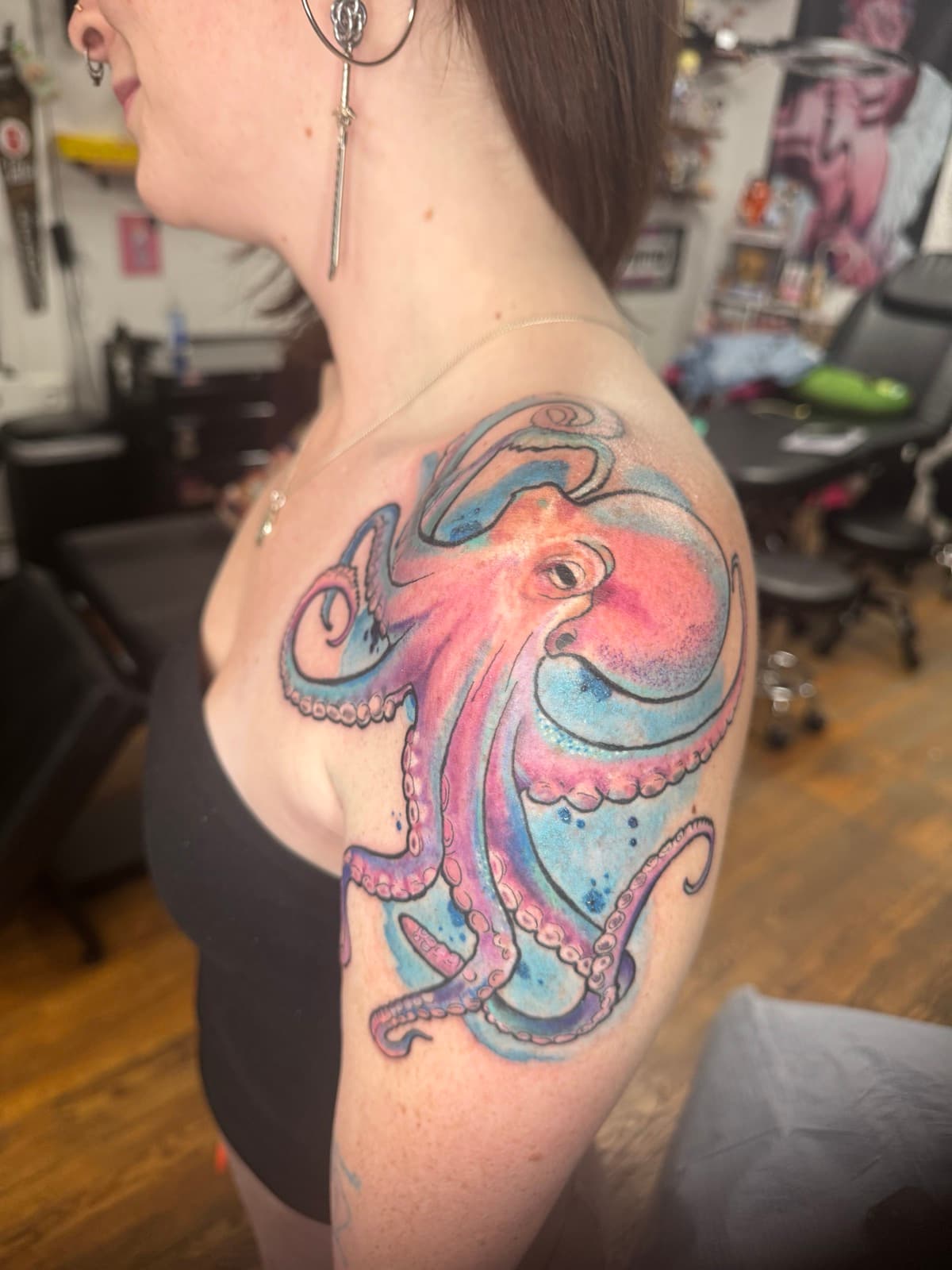 Large pink and coral watercolor octopus tattoo on shoulder — flowing tentacles wrapping over the chest with blue and teal watercolor splash.