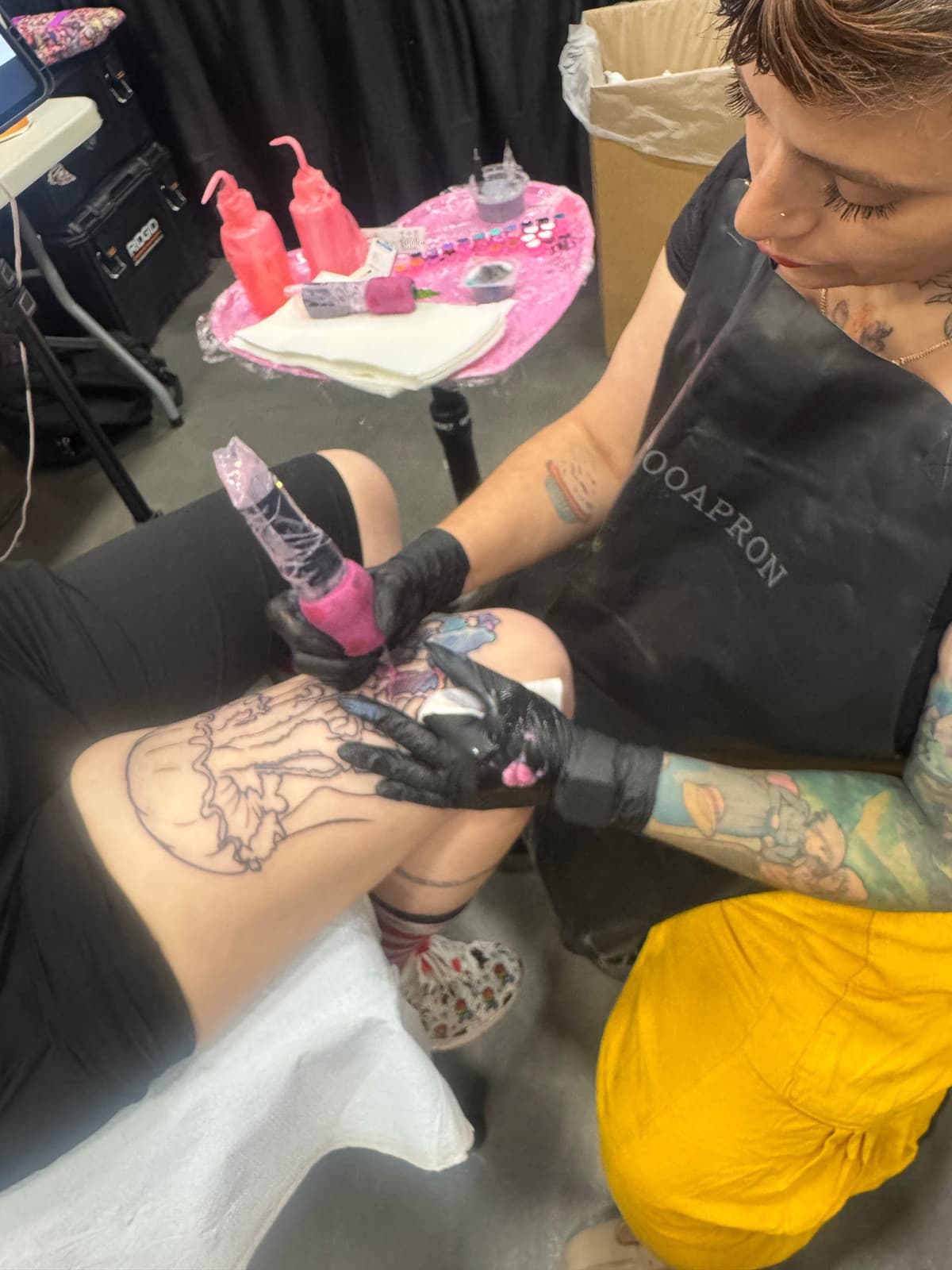 Paula working on a jellyfish line-work tattoo on a client's thigh, with pink wrap on the tattoo machine and a black "TOOAPRON" apron visible.