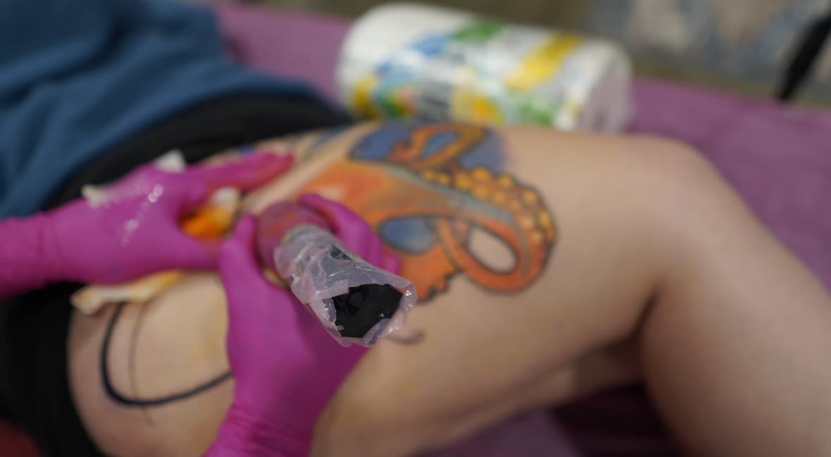 Close-up of a pink-gloved hand tattooing color into an orange watercolor octopus piece on a client's hip, with the tentacles and gold suckers visible.