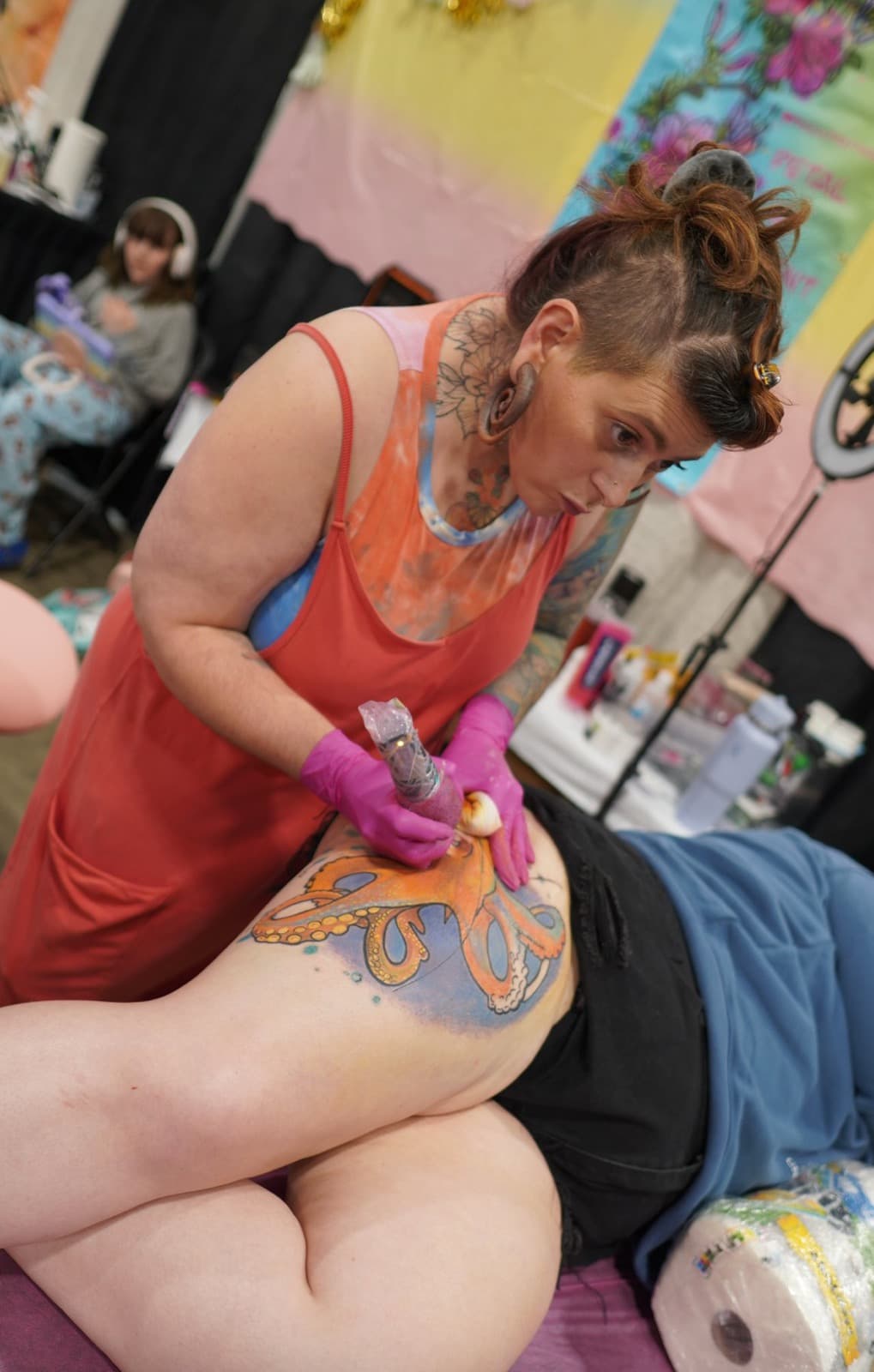 Paula working on the orange octopus tattoo at a convention, wearing a coral tank top and pink gloves, focused on the octopus tentacles and color work.