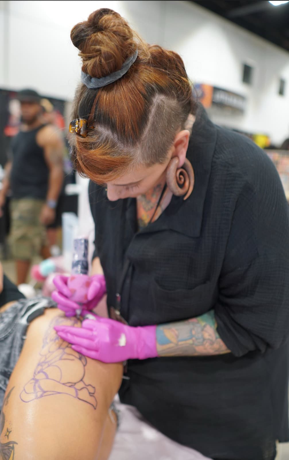 Paula tattooing at a convention with her hair in a top bun, shaved undercut, large round wood ear gauge, pink gloves, working purple stencil lines on a client's thigh.