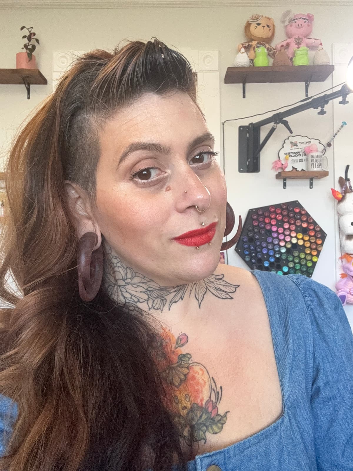 Paula, owner and artist at Petal & Pigment Tattoos, in her Cranford NJ studio — red lipstick, large wood ear gauges, septum piercing, visible neck and chest floral tattoos, with a rainbow paint palette wall behind her.