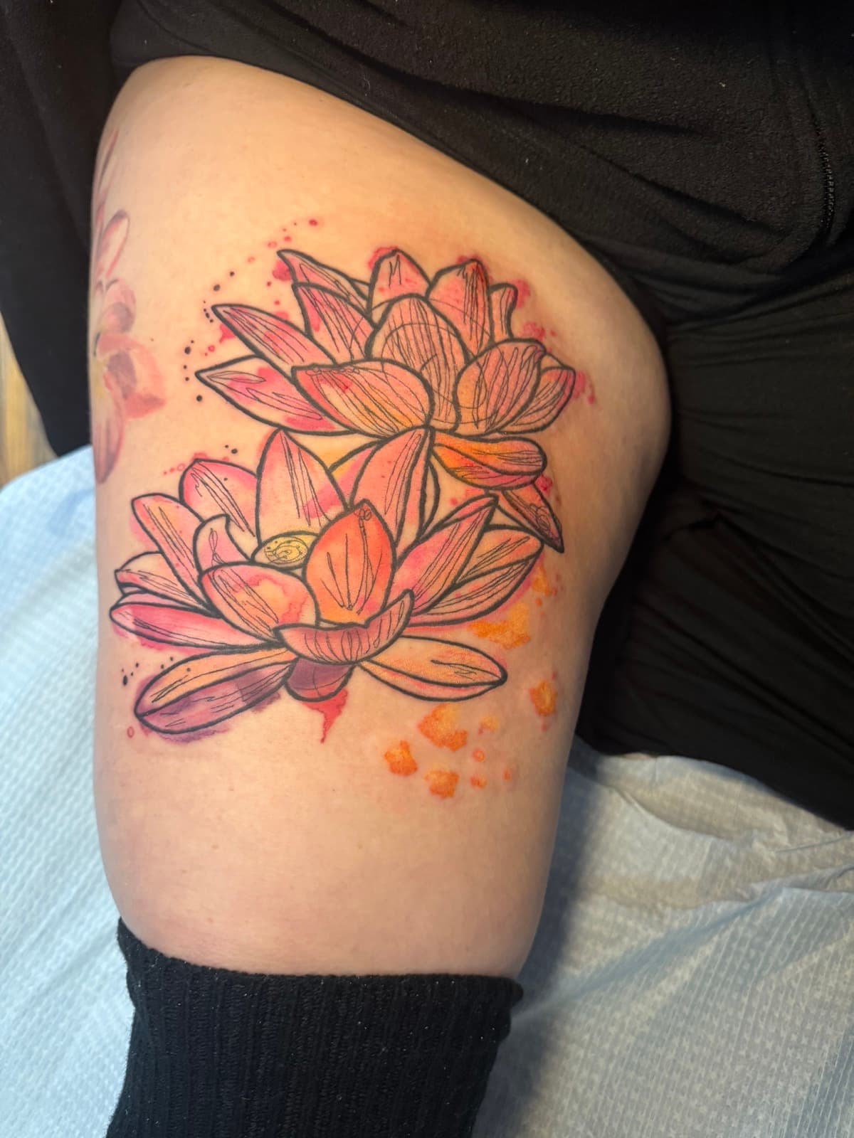 Watercolor lotus tattoo on thigh — two pink and coral lotus blossoms with line work and orange and red watercolor splash.